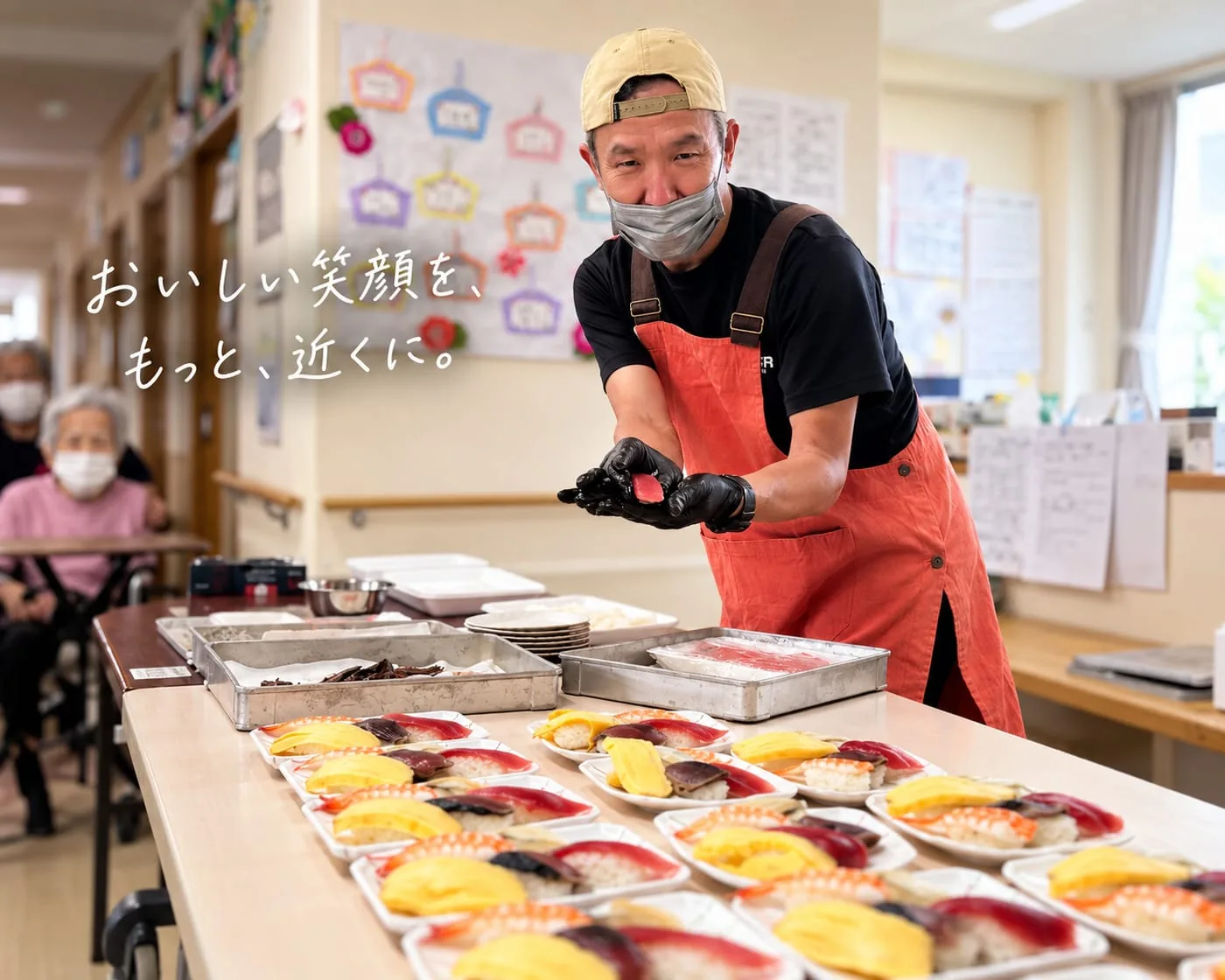 施設の食堂で、藤枝市場の店主が握った寿司を差し出している。手前には握り寿司の皿がずらり。写真には「おいしい笑顔を、もっと、近くに。」の手書き文字