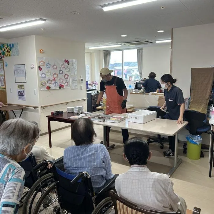 施設の食堂で、利用者さんたちがテーブルの向こうで寿司を用意する店主を見守っている